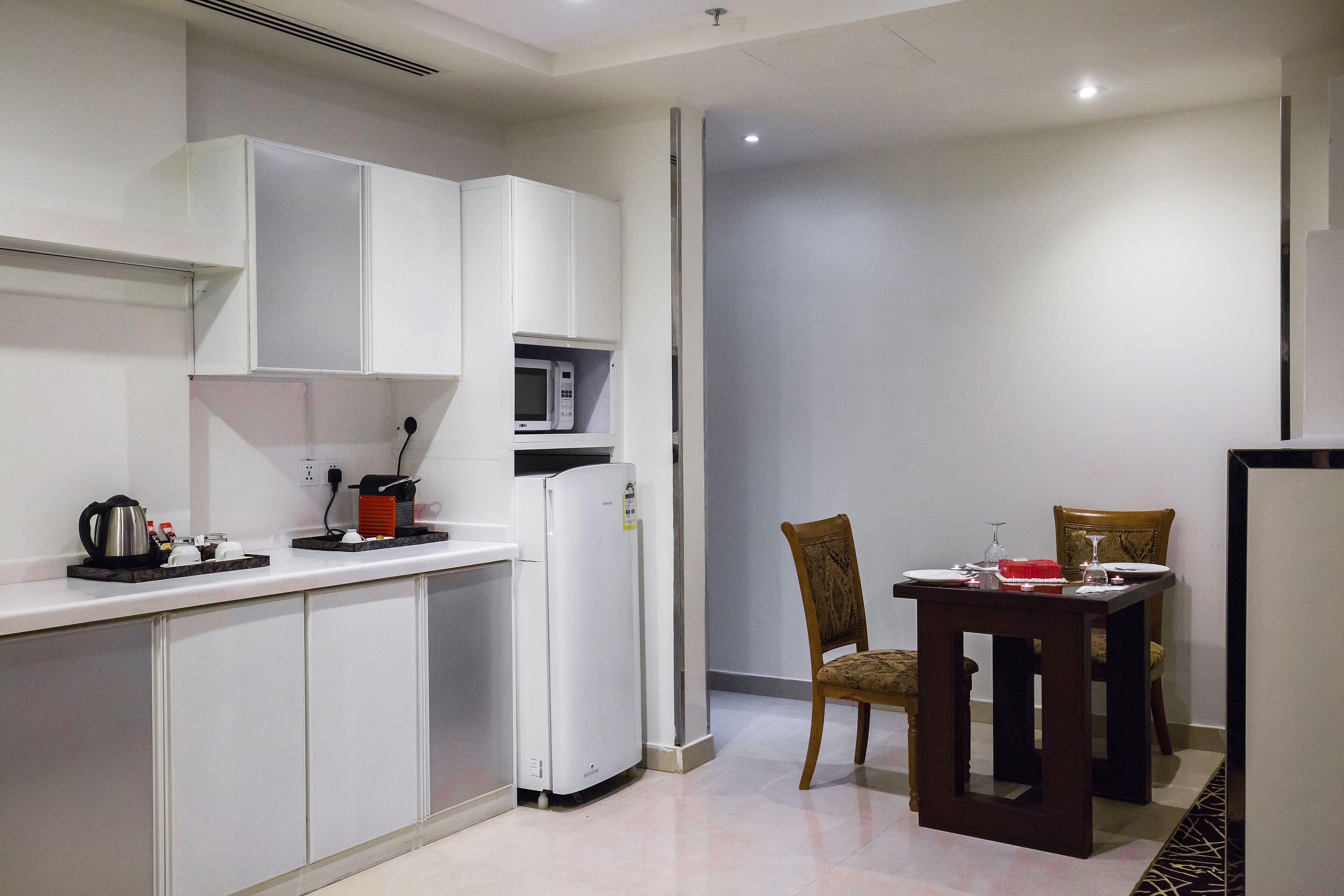 Una cucina compatta con mobili bianchi, un forno a microonde e un frigorifero. C'è un tavolo da pranzo con due sedie e una piccola lampada da tavolo.