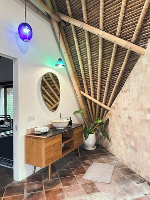 A bathroom with a wooden vanity, a round mirror, a large potted plant, and a bamboo ceiling.