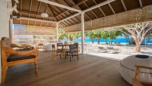Un espace extérieur aménagé, abrité et doté de mobilier en bois, comprenant une table et des chaises, qui offre une vue sur une plage avec des transats et sur l’océan.