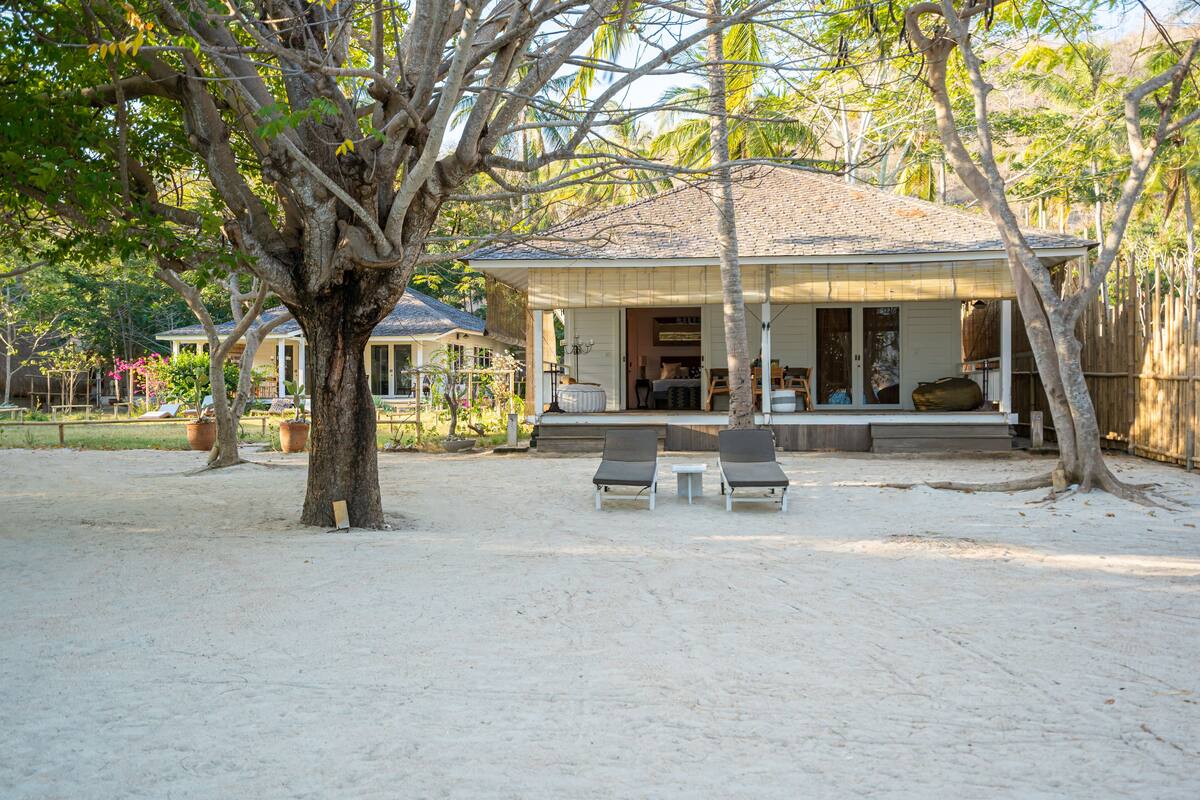 Een strandhuis met een overdekte veranda, twee lounge fauteuils en een grote boom op de voorgrond.