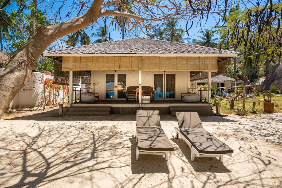 Een strandhuis met een overdekt terras, twee loungestoelen en een rieten dak.