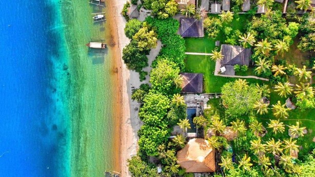 Vue aérienne d’une station balnéaire dotée de nombreux hébergements de type bungalow, entourés d’une végétation luxuriante et bordée par une mer turquoise aux eaux cristallines.