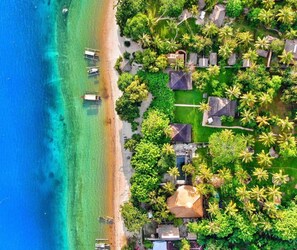 An aerial view of a coastal resort with multiple bungalow-style accommodations surrounded by lush greenery and a clear turquoise sea.