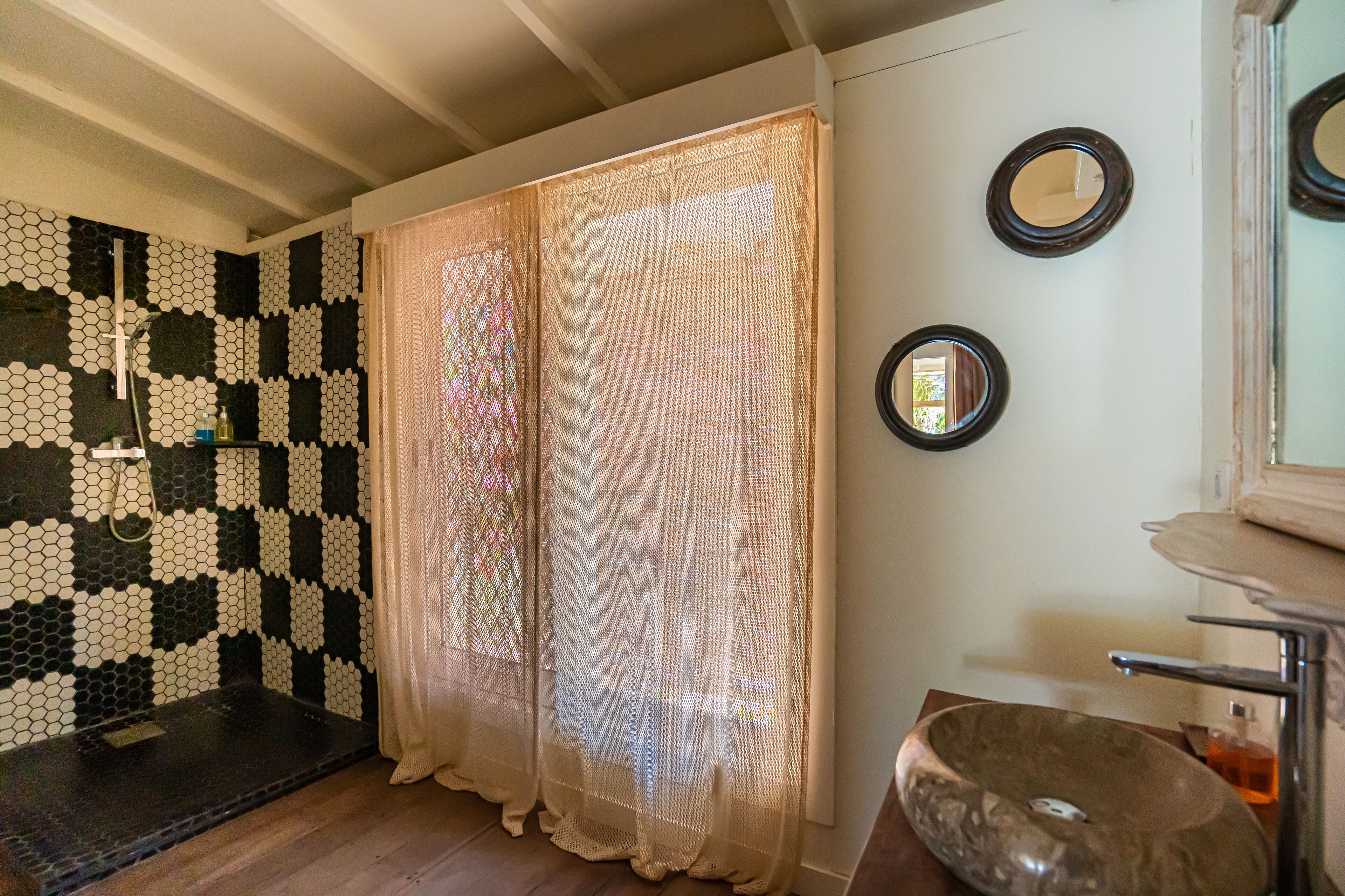A bathroom with a checkered mosaic wall, a large window with sheer curtains, a round mirror, and a vessel sink.