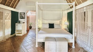 Une pièce spacieuse avec un plafond haut et un lit à baldaquin. Il y a un petit banc au pied du lit, ainsi qu’une table de chevet avec une lampe. La pièce est dotée d’un sol carrelé et d’une fenêtre avec des rideaux.
