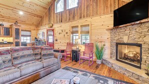 A log cabin interior with a stone fireplace, wooden floors, and a large window. - Hot Tub + Fire Pit: Dog-Friendly Cabin in Murphy! (Murphy)