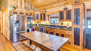A rustic wooden cabin interior with a large wooden table, built-in wooden cabinets, a stainless steel refrigerator, and a black cabinet with a window. - Hot Tub + Fire Pit: Dog-Friendly Cabin in Murphy! (Murphy)