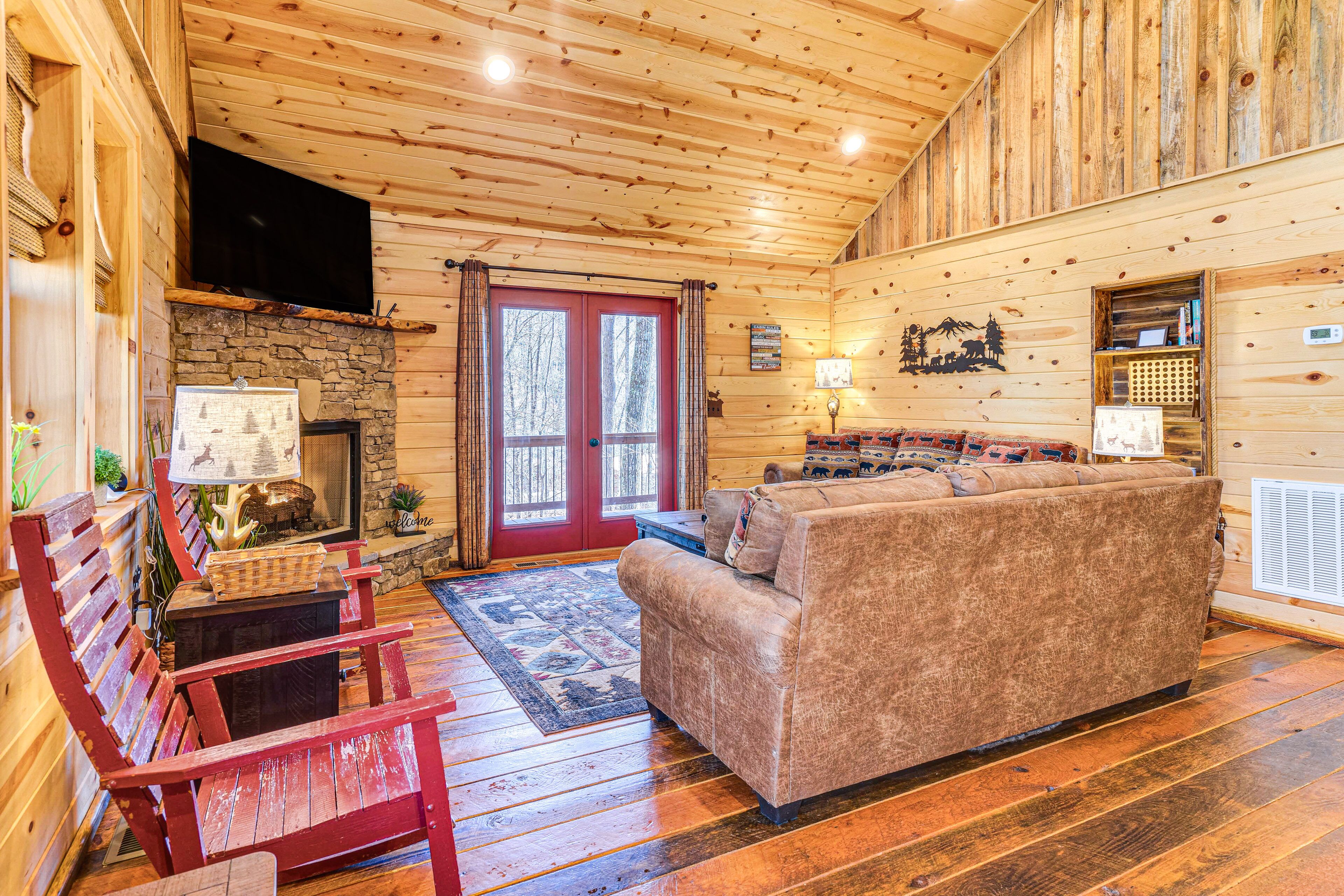 A log cabin interior with a stone fireplace, wooden floors, a red door, and a cozy seating area.