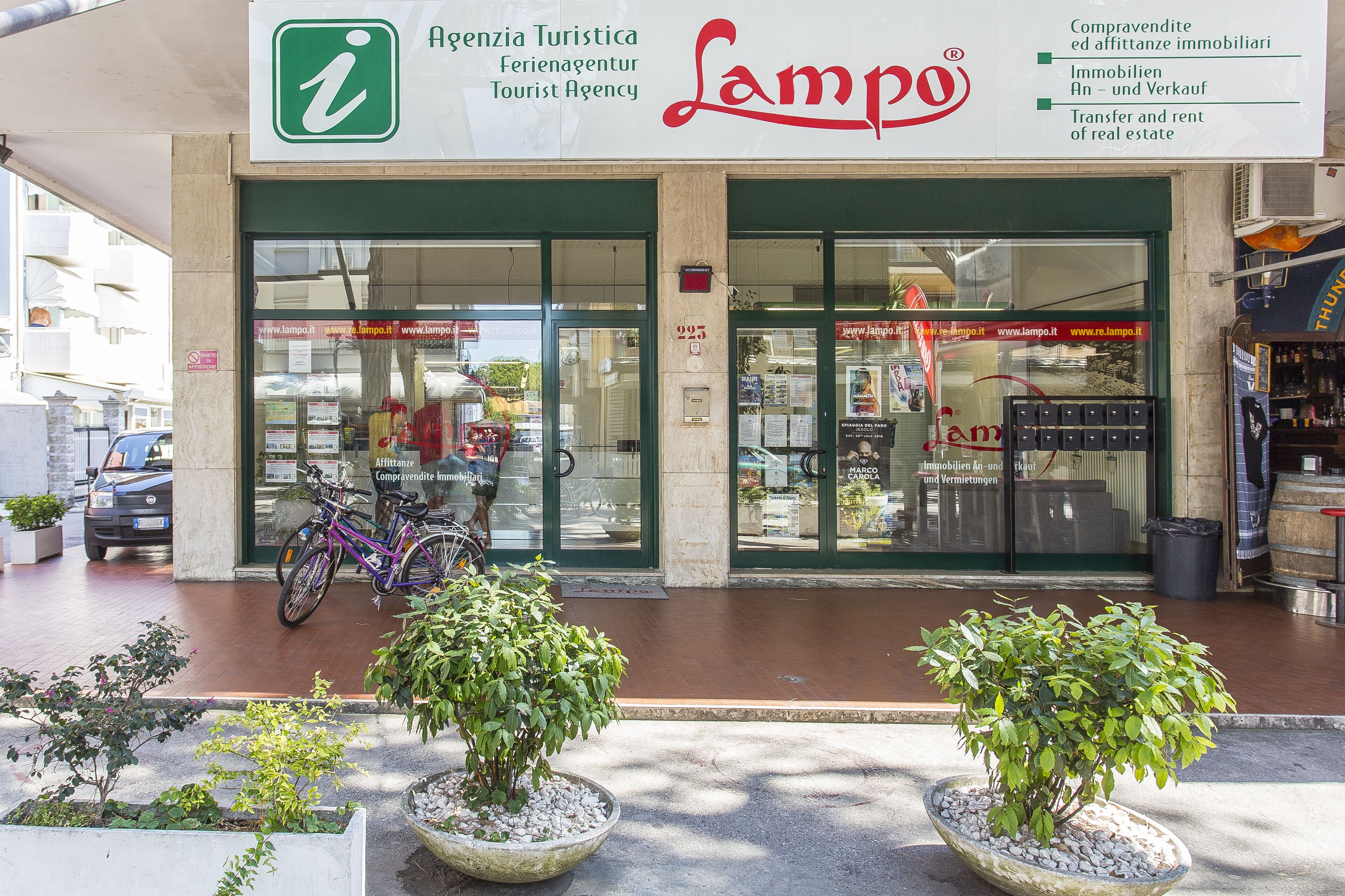 A tourist agency named 'Lampo' with a green and white sign, a bicycle parked outside, and potted plants in front.