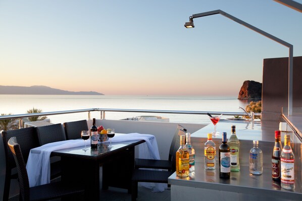 A balcony with a table set for dining, a bar area with various bottles, and a view of the sea and distant land.