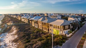 A coastal residential area with houses featuring large windows and balconies, a beach in the background, and a road curving along the coast.