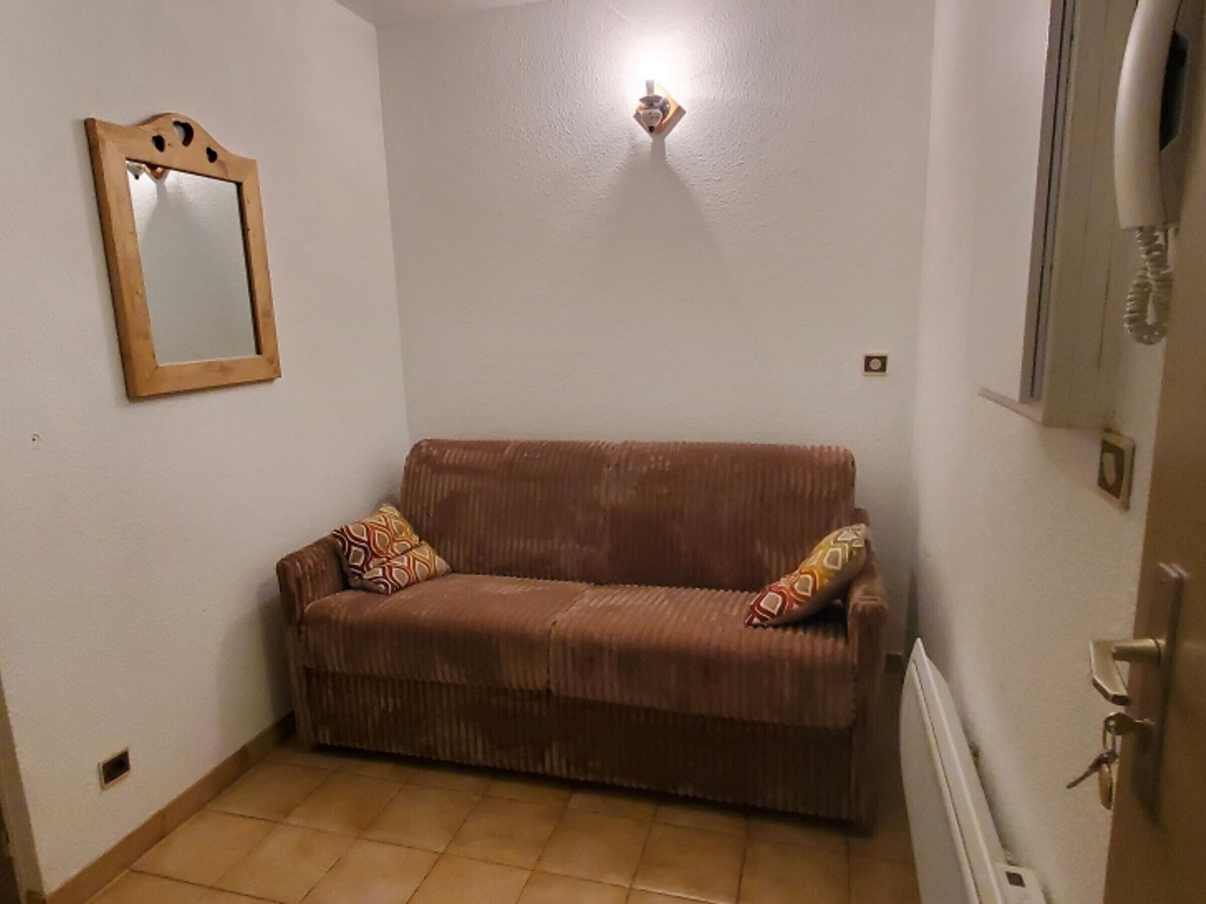 A small, cozy room with a brown sofa, a wooden-framed mirror, and a wall-mounted light fixture.