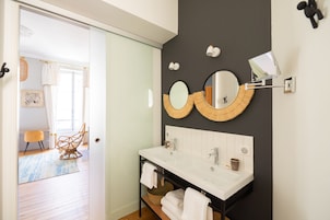 A modern bathroom with a double sink, two round mirrors, and a large mirror above the sink. There is a wicker basket under the sink and a towel rack with towels.