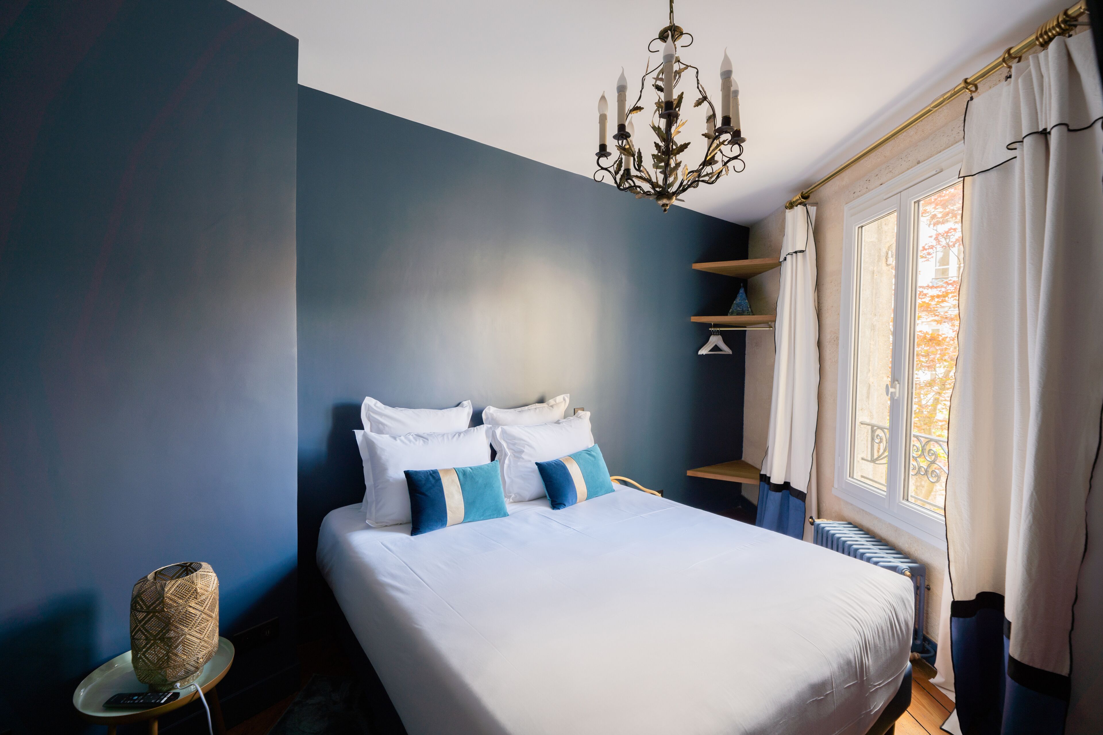 Un lit bien fait, avec du linge de lit blanc et des coussins bleus, un lustre, une table de chevet avec une lampe et une fenêtre avec des rideaux.