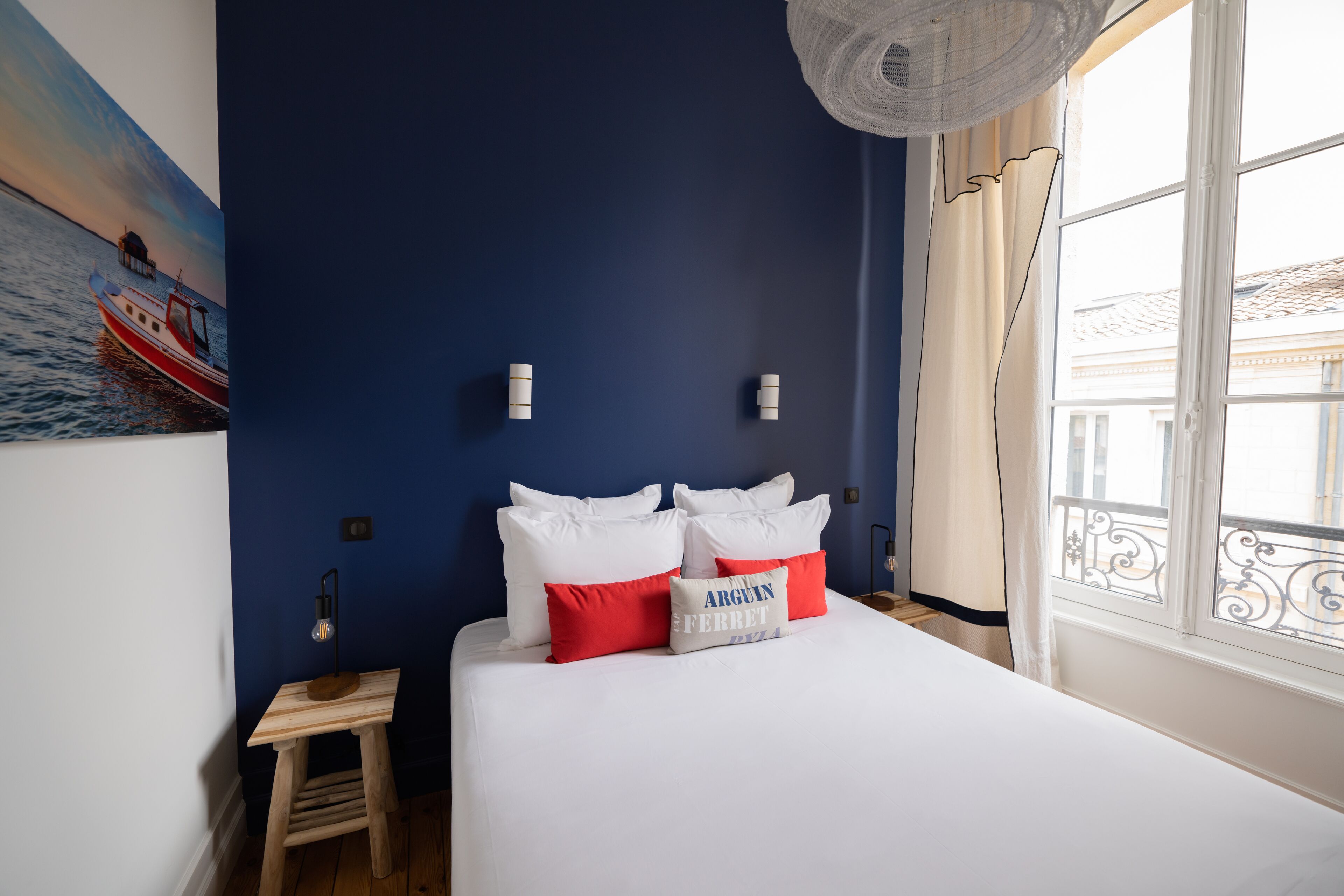 Un dormitorio con cama, almohadas, una mesita de noche, un cuadro y una ventana con vistas a edificios.