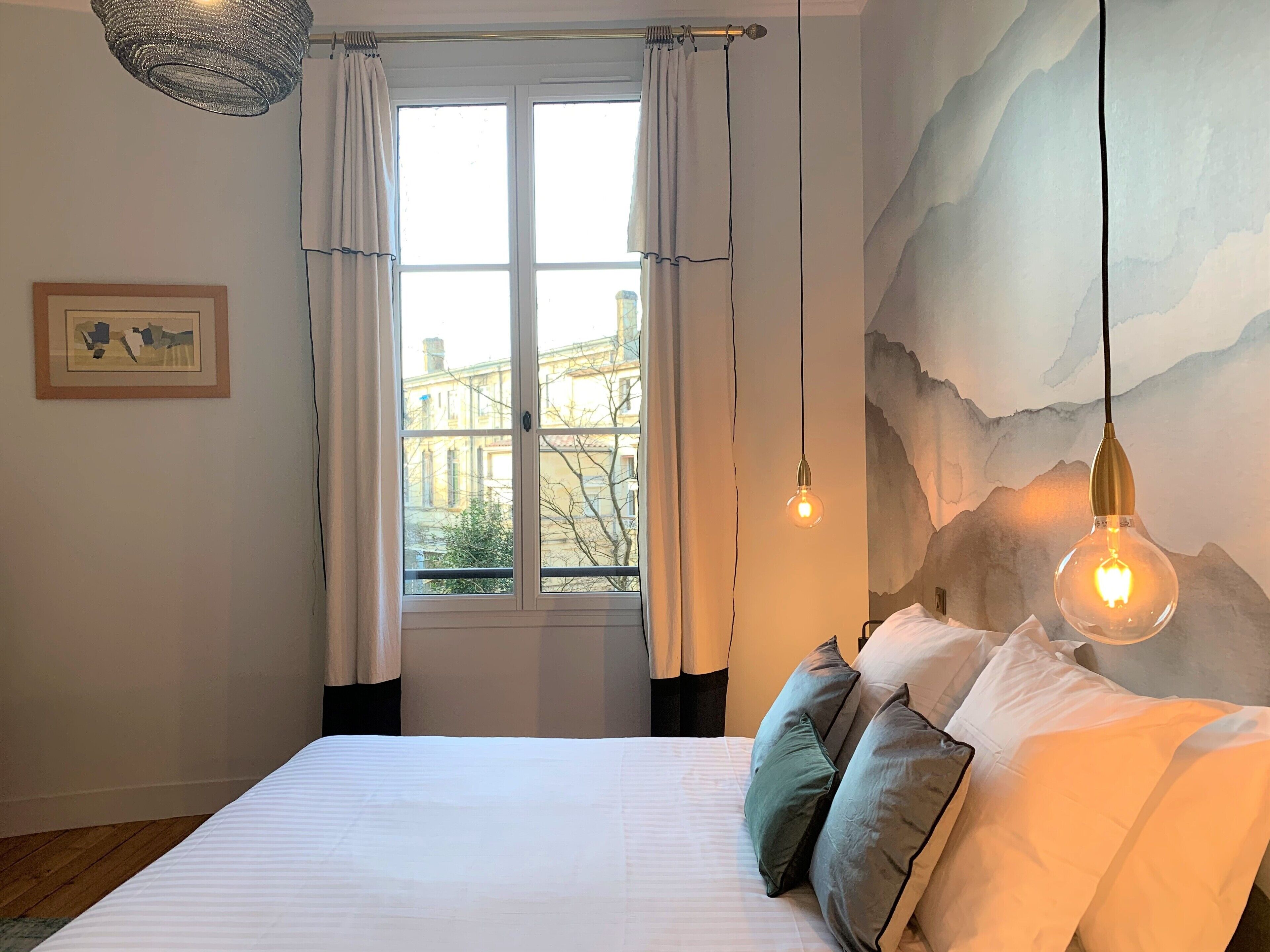 Une chambre avec un grand lit, du linge de lit blanc, une vue par la fenêtre et un luminaire fixé au mur.