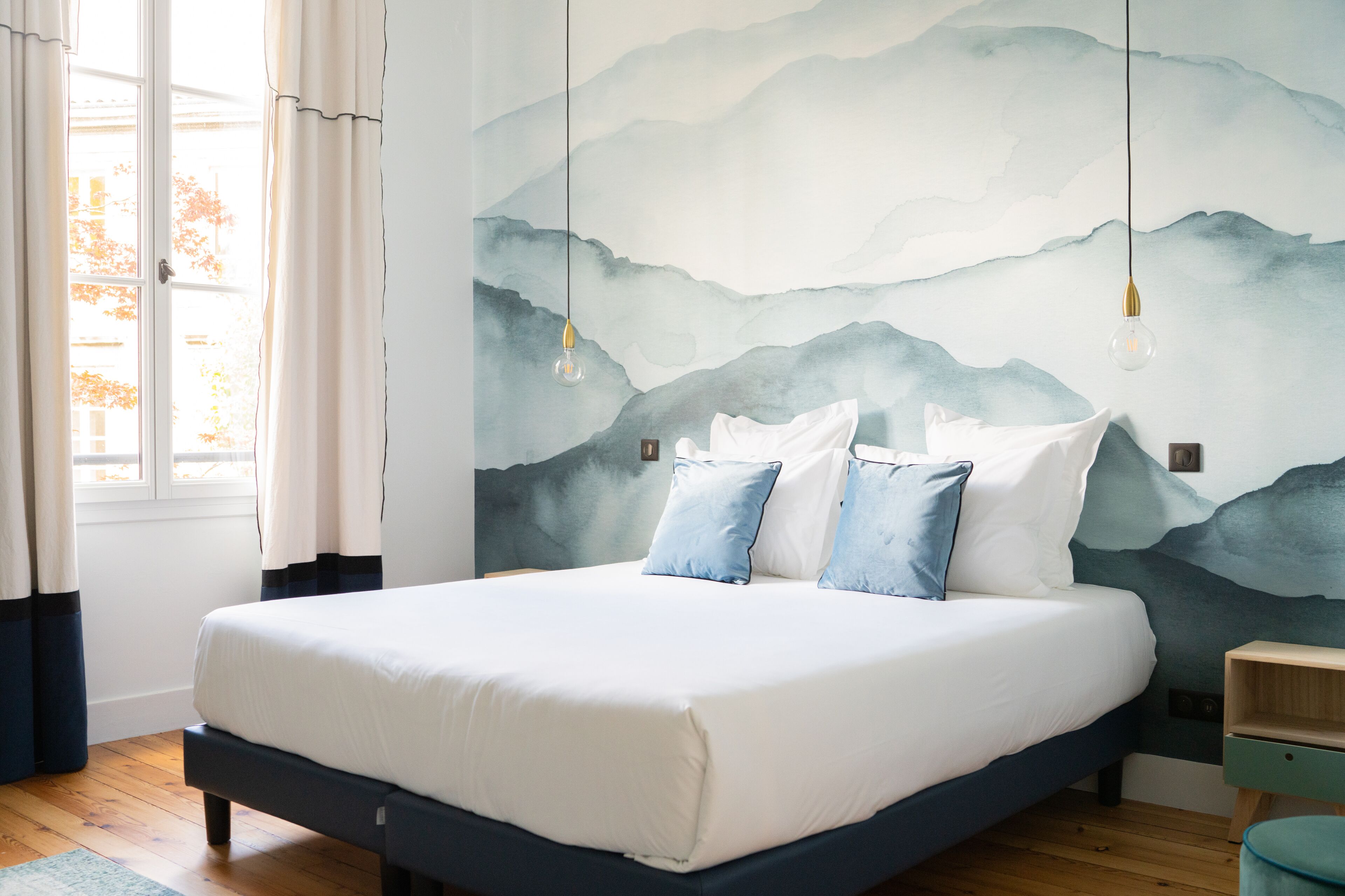 A neatly made bed with white and blue bedding, a wooden floor, a wall with a mountain landscape mural, and a window with curtains.
