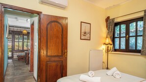 Una habitación de hotel con puerta de madera, una cama con ropa blanca, un cuadro de un velero, una ventana con cortinas y un radiador.
