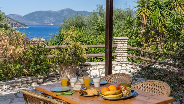 Una mesa de madera con platos, vasos y frutas, ubicada al aire libre con vistas al mar y las montañas.