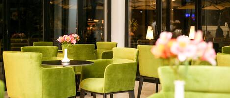 A seating area with green upholstered chairs and tables, a white vase with flowers, and a view through large glass windows.