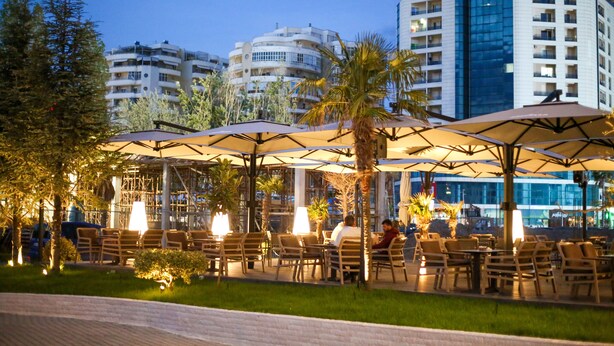 An outdoor seating area with umbrellas and chairs, surrounded by greenery and buildings in the background. - Hotel Regina Garden Vlore (Vlorë)