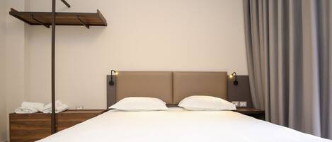 Une chambre d’hôtel avec un grand lit, une table de chevet en bois, une étagère fixée au mur et une petite table de chevet avec une lampe.