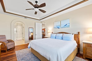 Une chambre avec un grand lit, une tête de lit en bois, un ventilateur de plafond, une chaise en bois, une douche en verre et une porte donnant sur une autre pièce.