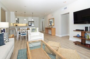 A modern living room with a sofa, coffee table, and TV stand. There is a dining area with a table and chairs, and a kitchen with cabinets and a sink.