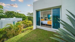 Habitación de hotel moderna con un amplio ventanal corredero, una cama con manta amarilla y vistas a la vegetación circundante.