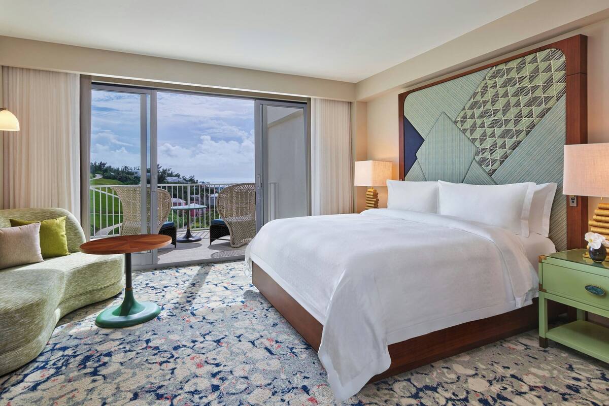 A hotel room with a large bed, a sofa, a small table, and a balcony with a view of the landscape.