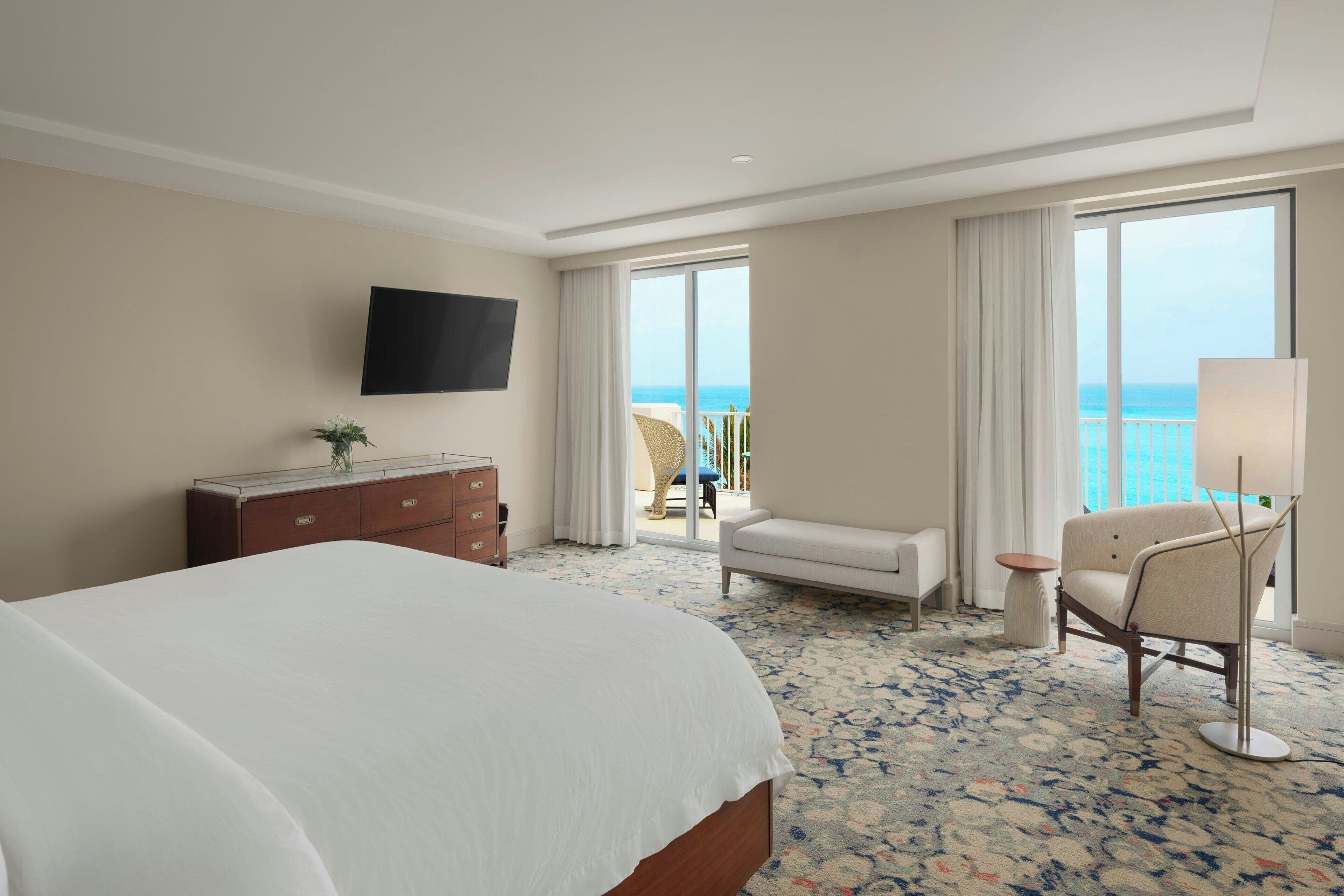A hotel room with a large bed, a flat-screen TV mounted on the wall, a sofa, a chair, and a small table. The room has a large window with a view of the ocean.