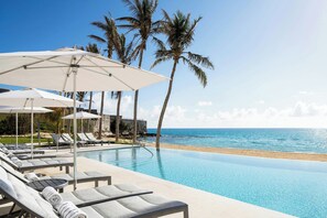 A poolside area with lounge chairs and umbrellas, a beach view, and palm trees.