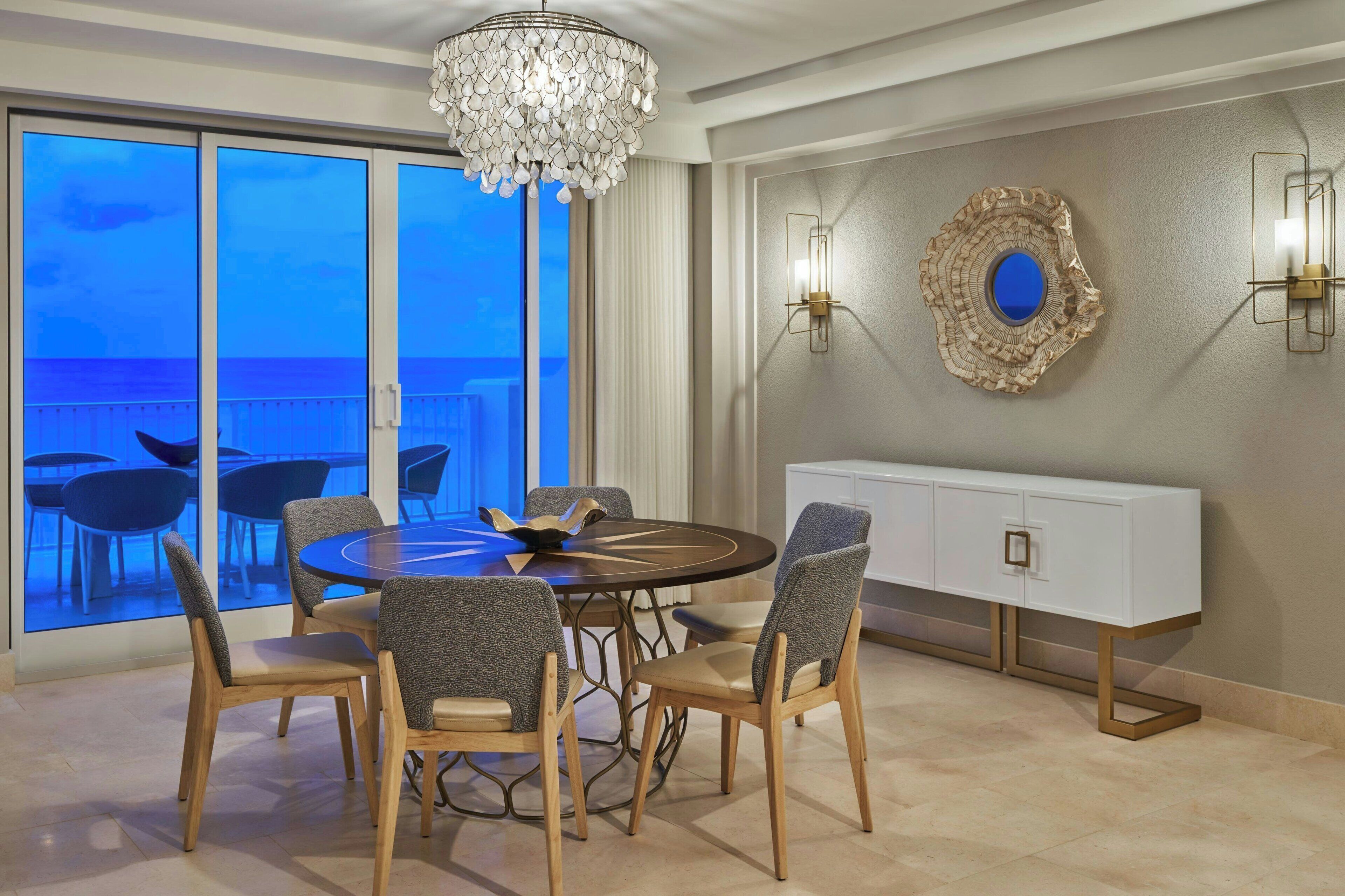 Un espace repas comprenant une table ronde et des chaises, un lustre, une console blanche et une vue dégagée sur l’extérieur grâce à de grandes fenêtres.