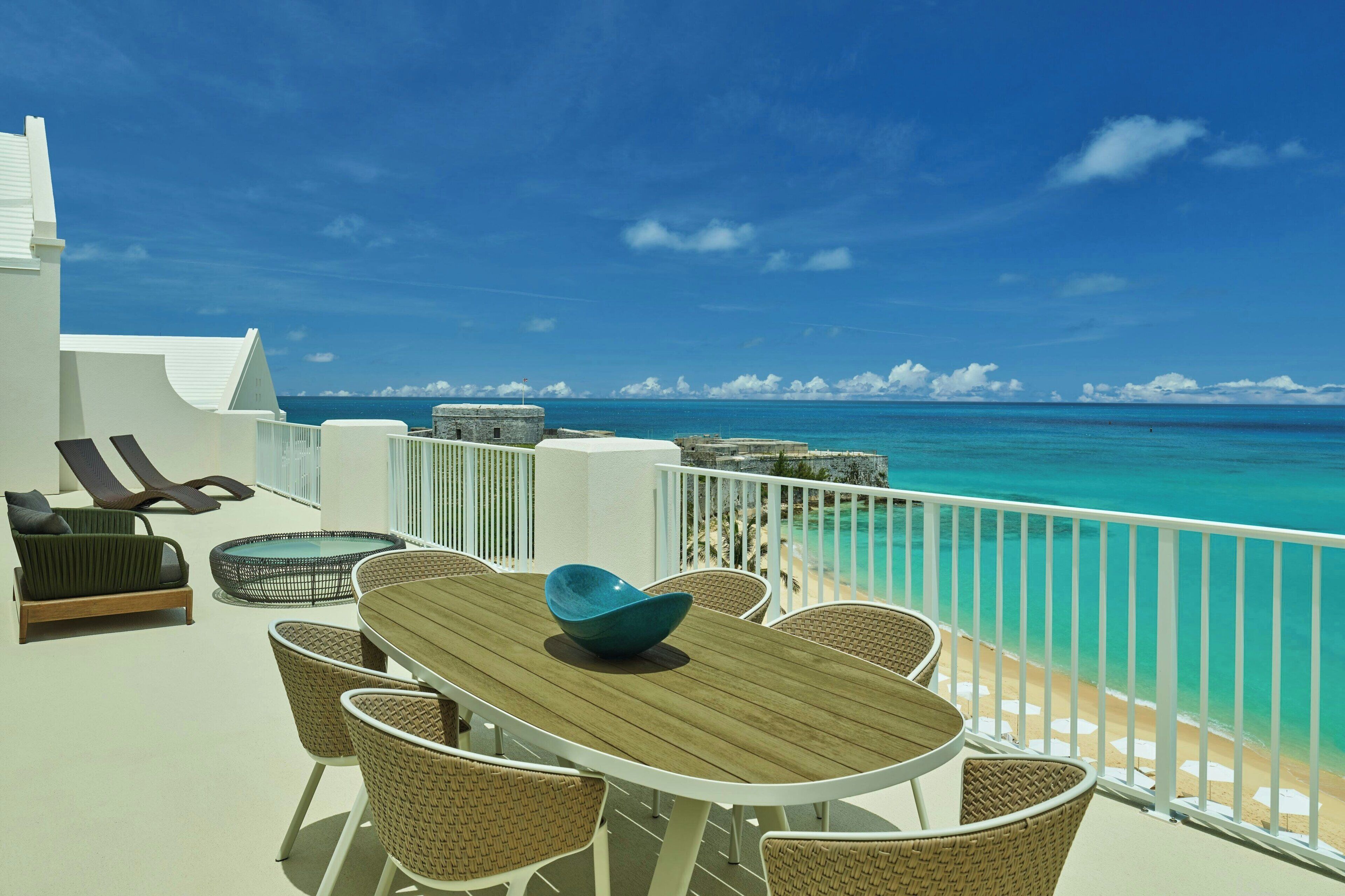 Un balcon doté d’une table en bois et de chaises en osier, offrant une vue sur une plage aux eaux turquoise et limpides, ainsi que sur une balustrade blanche.