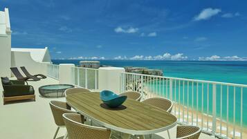 Un balcon doté d’une table en bois et de chaises en osier, offrant une vue sur une plage aux eaux turquoise et limpides, ainsi que sur une balustrade blanche.