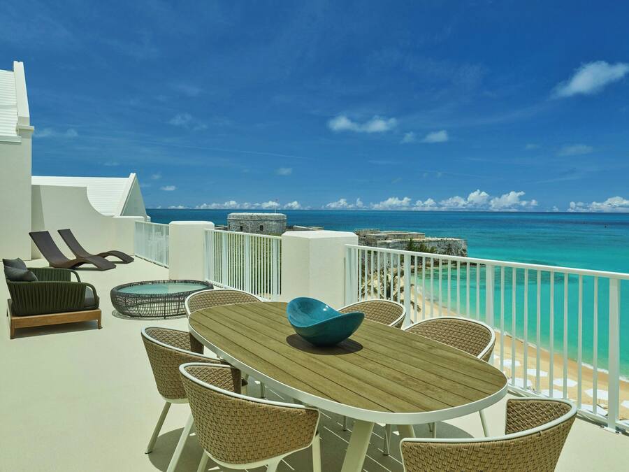 Un balcon doté d’une table en bois et de chaises en osier, offrant une vue sur une plage aux eaux turquoise et limpides, ainsi que sur une balustrade blanche.