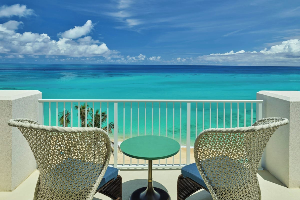 A balcony with wicker chairs and a small table, overlooking a turquoise sea and a clear sky.