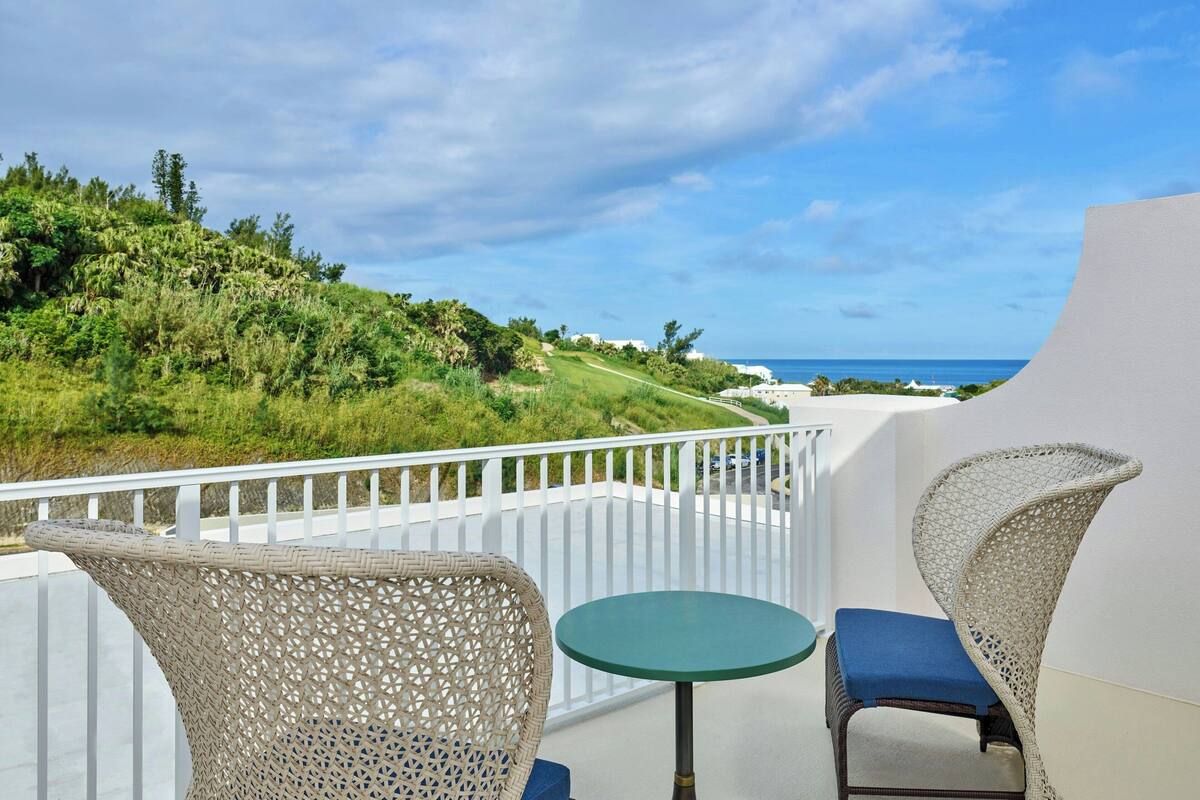 A balcony with wicker chairs and a small table, overlooking a lush green landscape and the ocean.