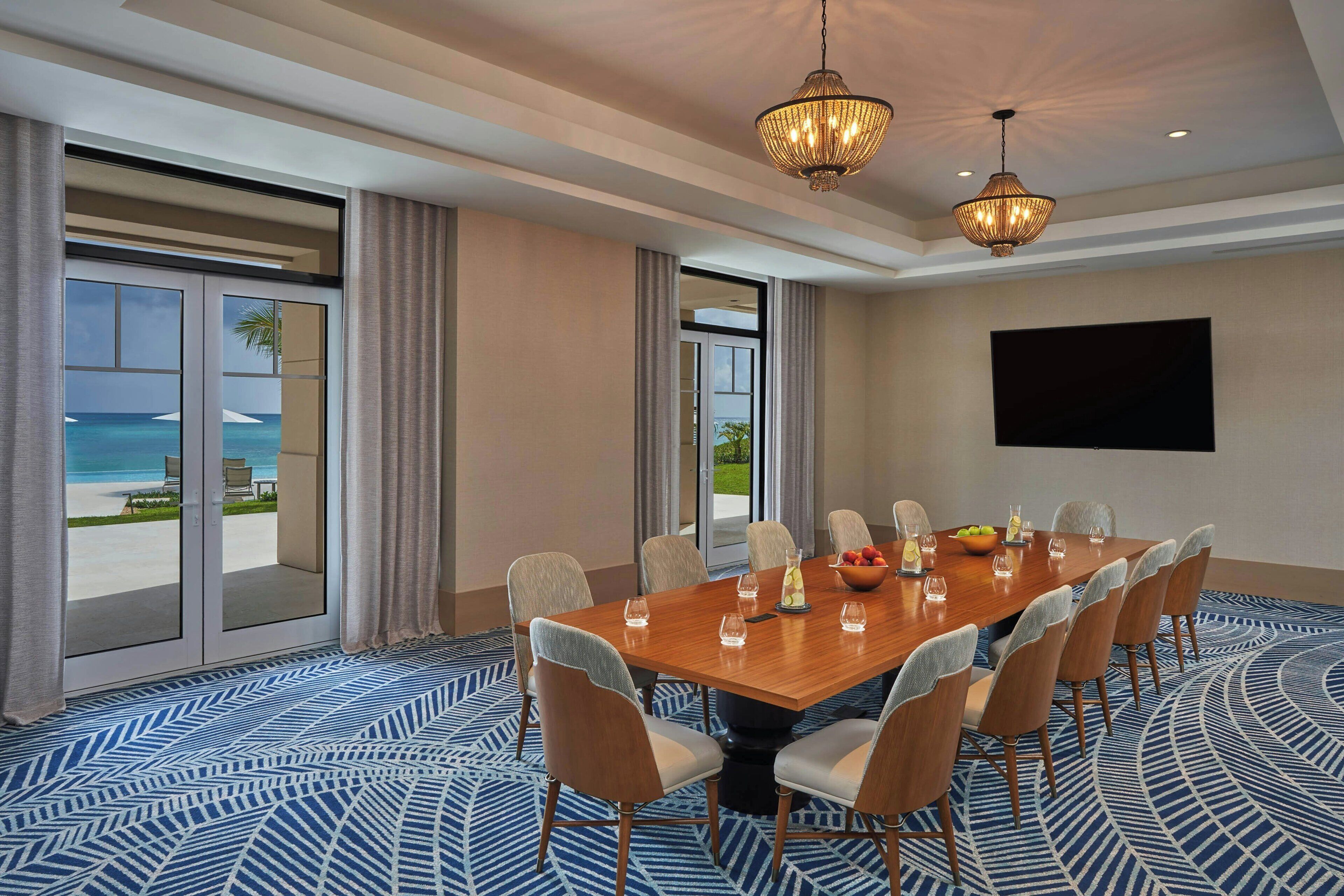A conference room with a large table, chairs, a chandelier, and a large window with a view of the beach.