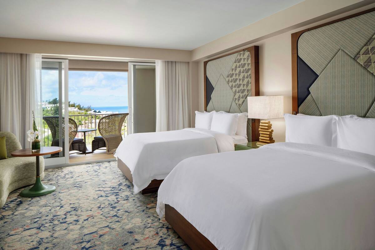 A hotel room with two beds, a balcony with outdoor seating, and a view of the ocean.