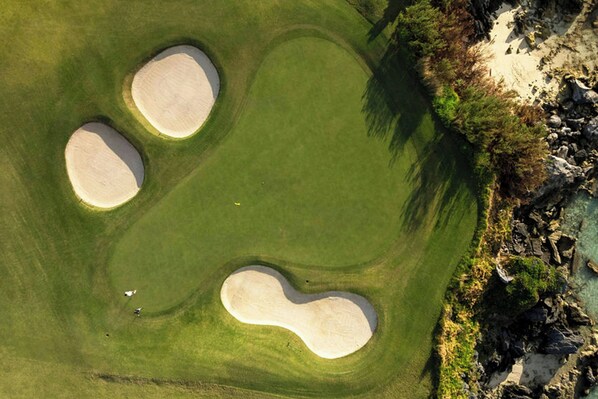 An aerial view of a golf course with three sand bunkers and a rocky shoreline.