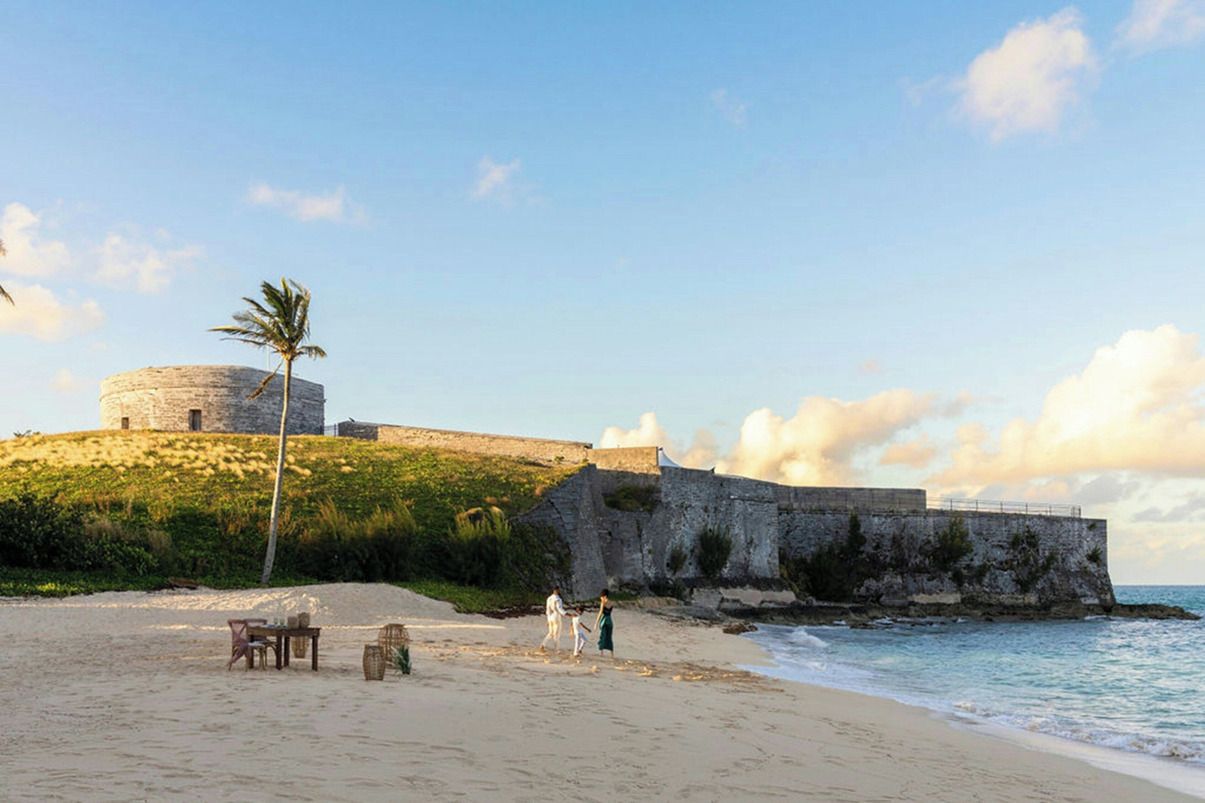 A beach with a stone fortification, a palm tree, and a table with chairs.