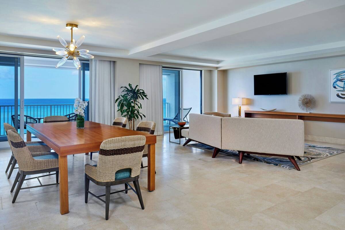 A spacious living area with a wooden dining table, wicker chairs, a sofa, and a TV mounted on the wall.