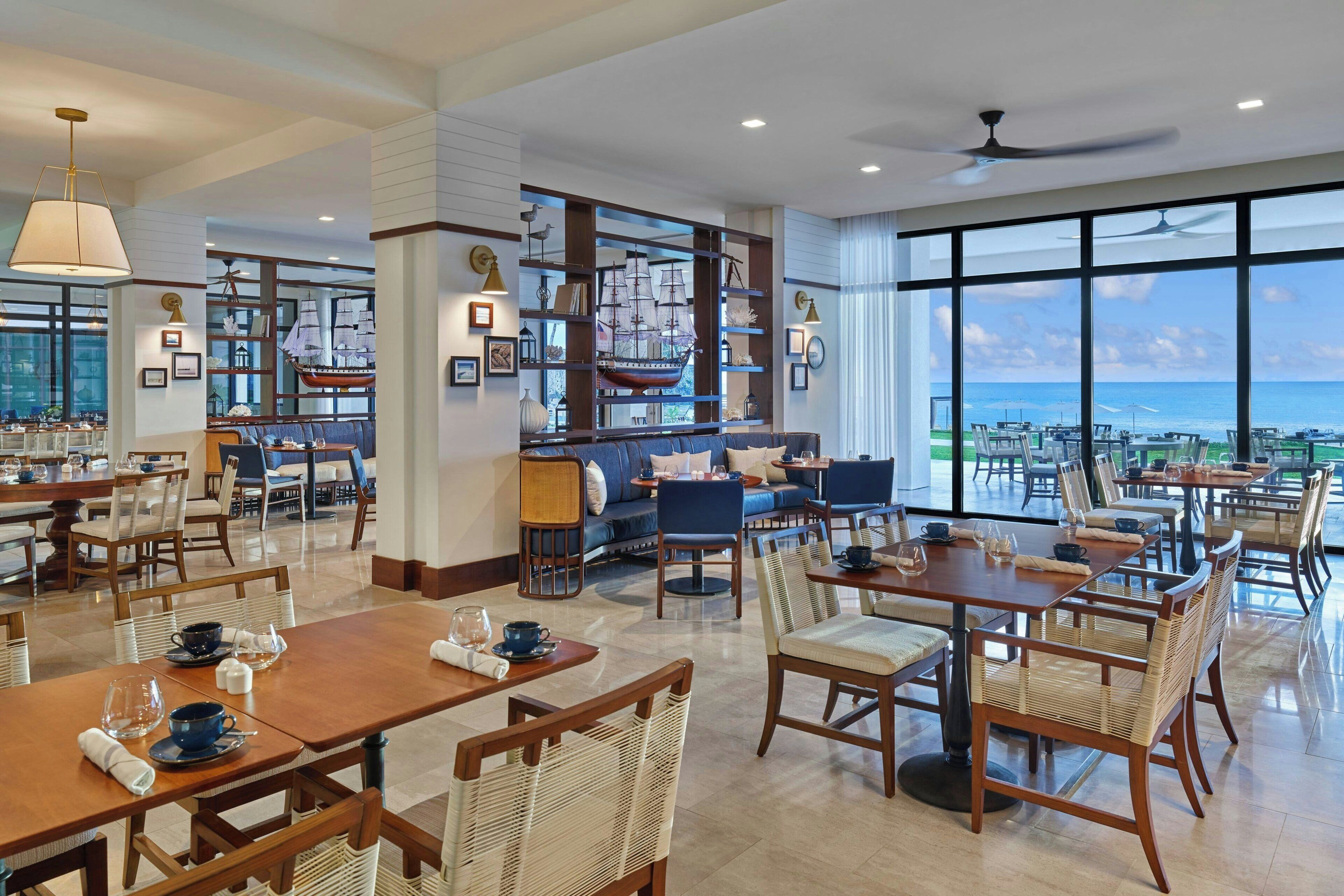 Un comedor amplio con mesas y sillas altas, una zona de bar con un gran modelo de barco, y vistas al océano a través de ventanas panorámicas.