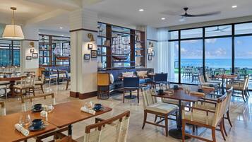 Un comedor amplio con mesas y sillas altas, una zona de bar con un gran modelo de barco, y vistas al océano a través de ventanas panorámicas.