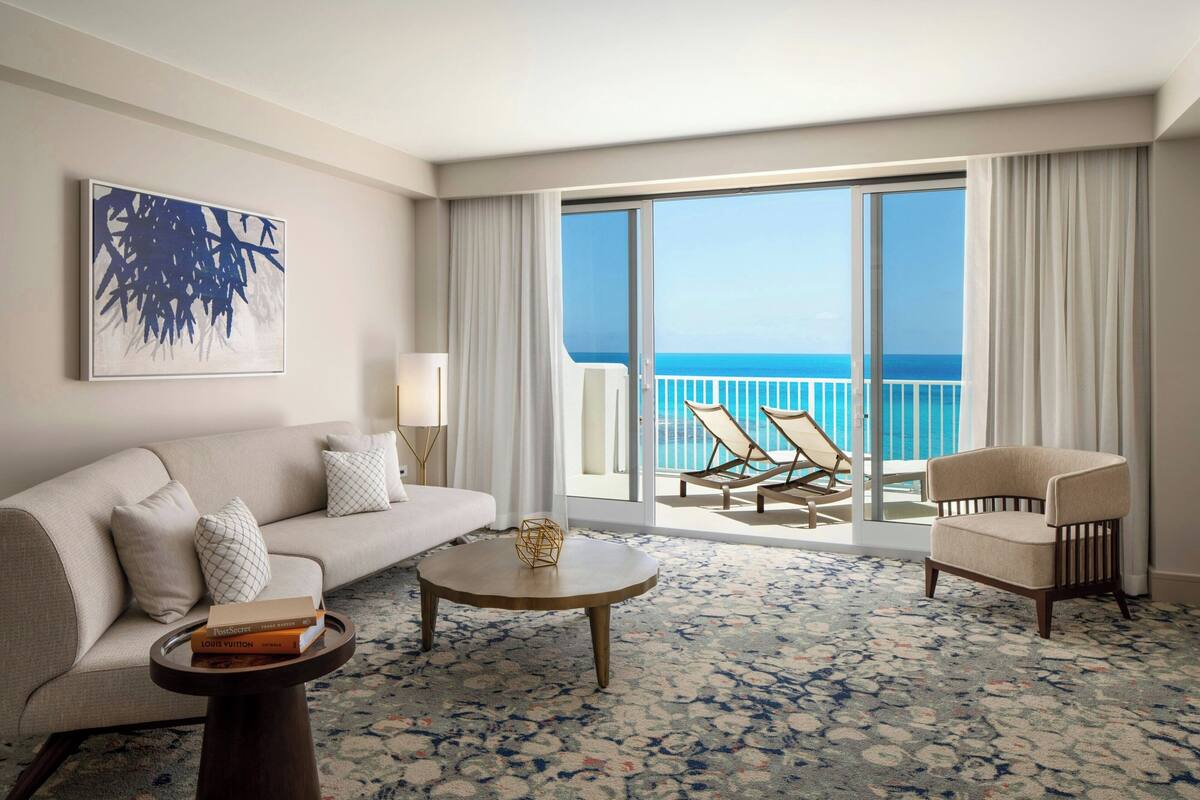 A hotel room with a sofa, armchair, round coffee table, and a view of the ocean through large sliding doors.
