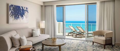 A hotel room with a sofa, armchair, round coffee table, and a view of the ocean through large sliding doors.