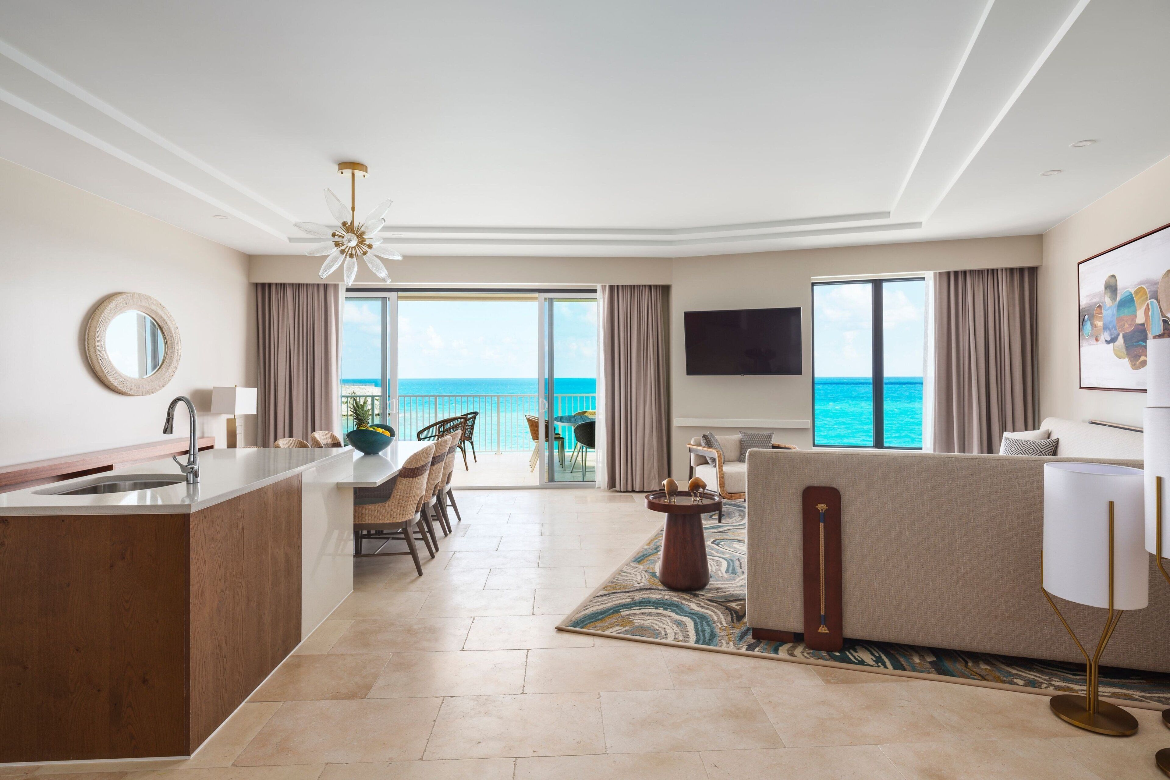 A modern hotel room with a kitchen area, dining space, and a large balcony with a view of the ocean.