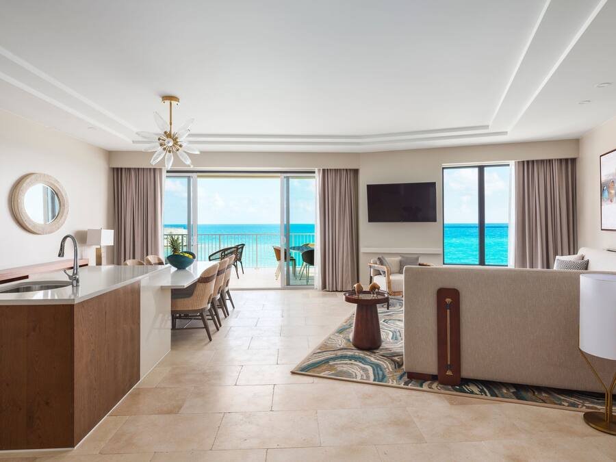 A modern hotel room with a kitchen area, dining space, and a large balcony with a view of the ocean.