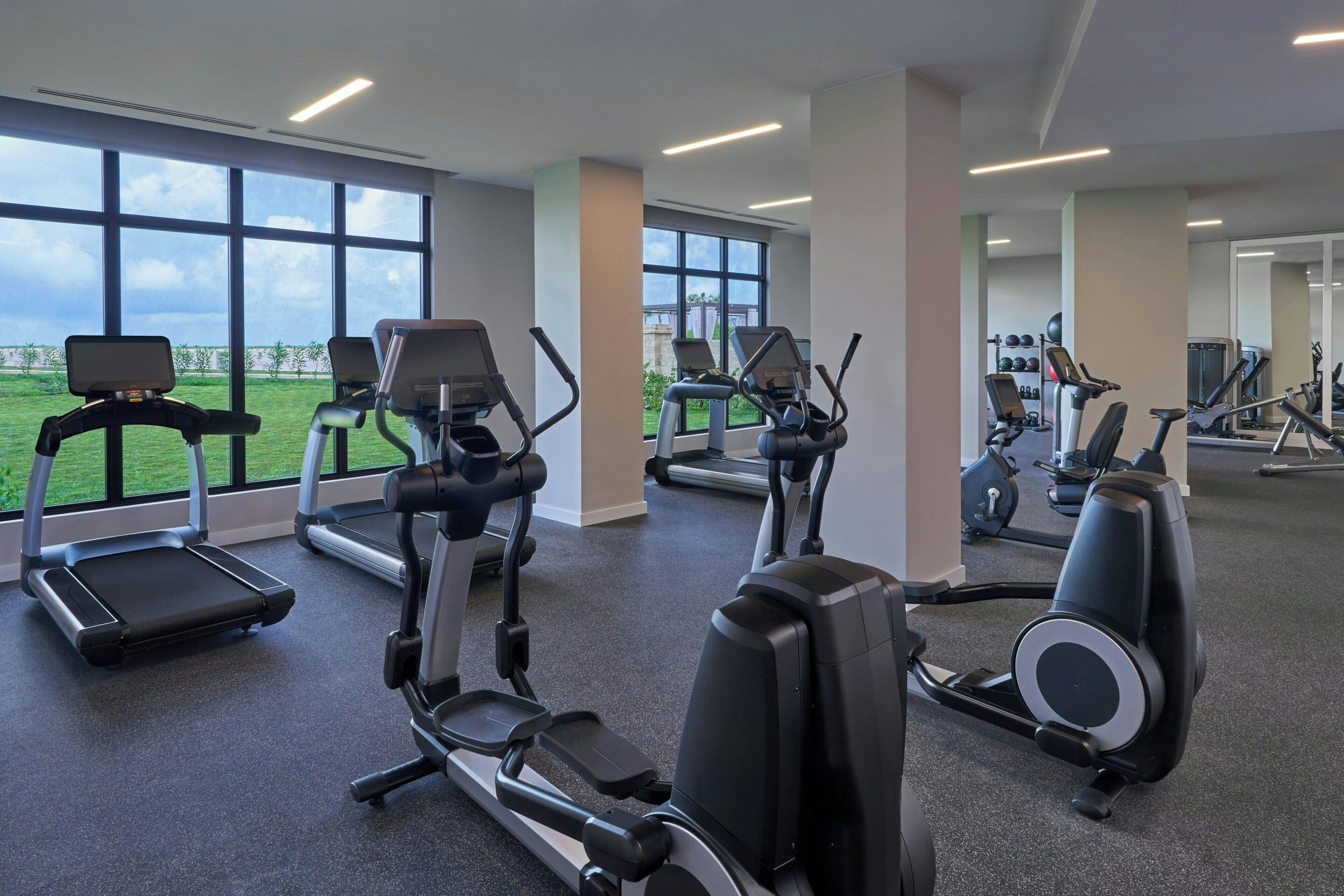 Une salle de sport équipée de tapis de course, de vélos elliptiques et offrant une vue sur un champ verdoyant grâce à de grandes fenêtres.